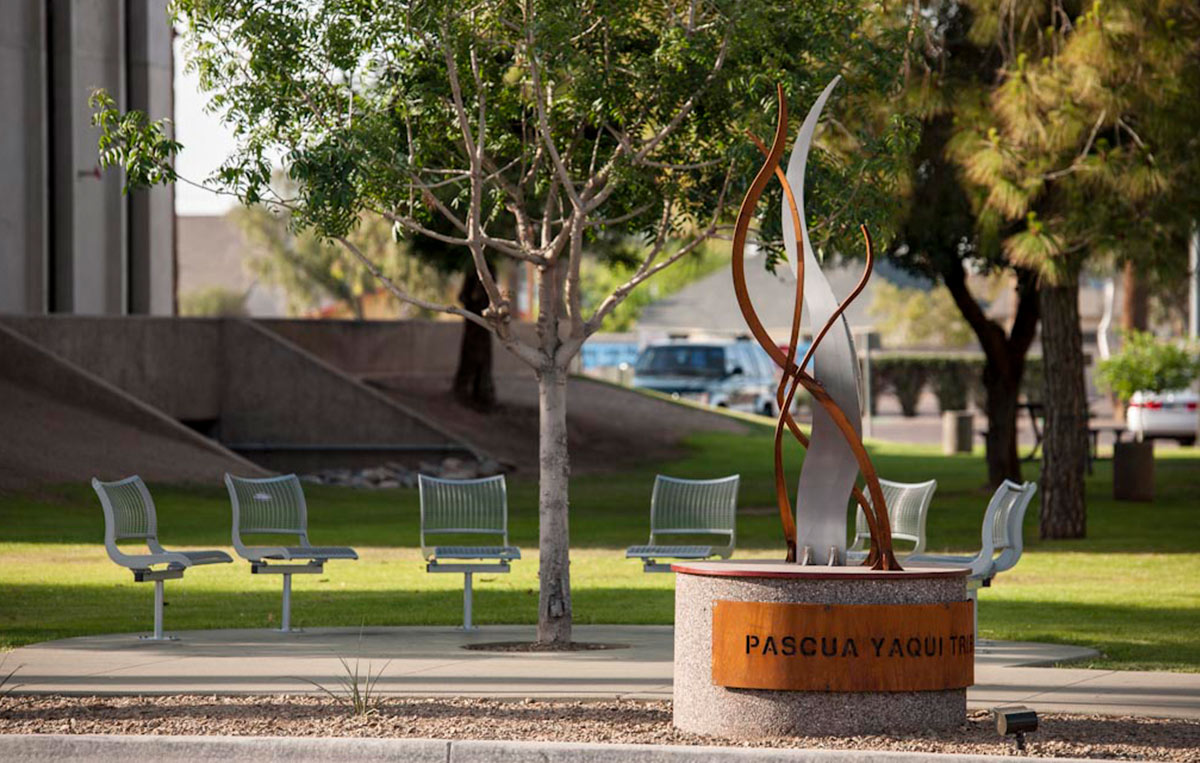 Photo: a sculpture in a streetscape. Undulating metal ribbons rise like flames above a heavy cylindrical concrete base. On the base is a metal place with the words cut out, "Pascua Yaqui Tribe."