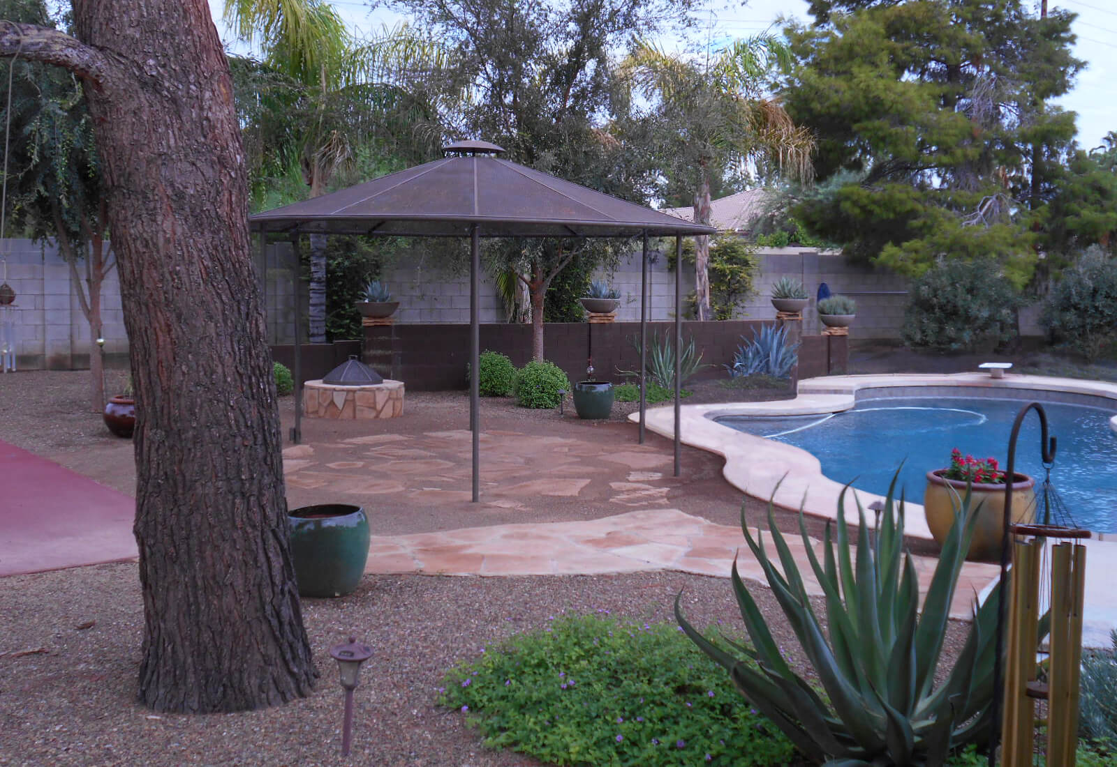 Photo of a backyard with a flagstone paver patio under a round metal shade structure. Next to the patio is a bean-shaped pool. The yard is shaded by pine trees and palm trees and is filled with flowering shrubs and agaves.