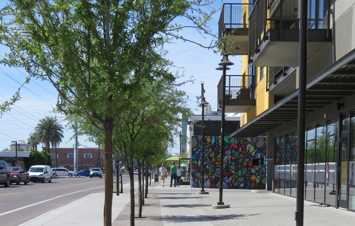 Photo of Roosevelt Row streetscape. Oaks provide shade along the sidewalk in front of a multi-use building with an abstract geometric mural on one wall.