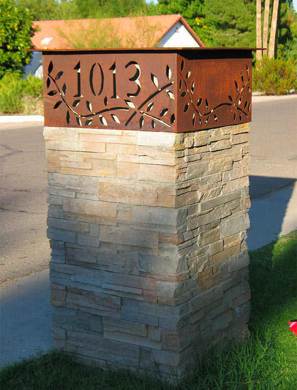 Photo of a custom-designed residential mailbox. The base is a thick column of pale stacked stone. The top is a rusted metal sheath over the mailbox itself, laser-cut with the house number and an abstracted leaf design.