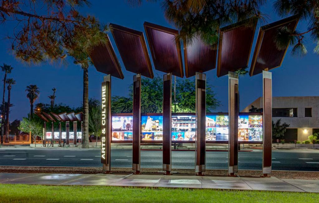 Photo of an artistic bus stop at night. Wide, flat, rectangular metal columns support rectangular panels that are set at different angles.
