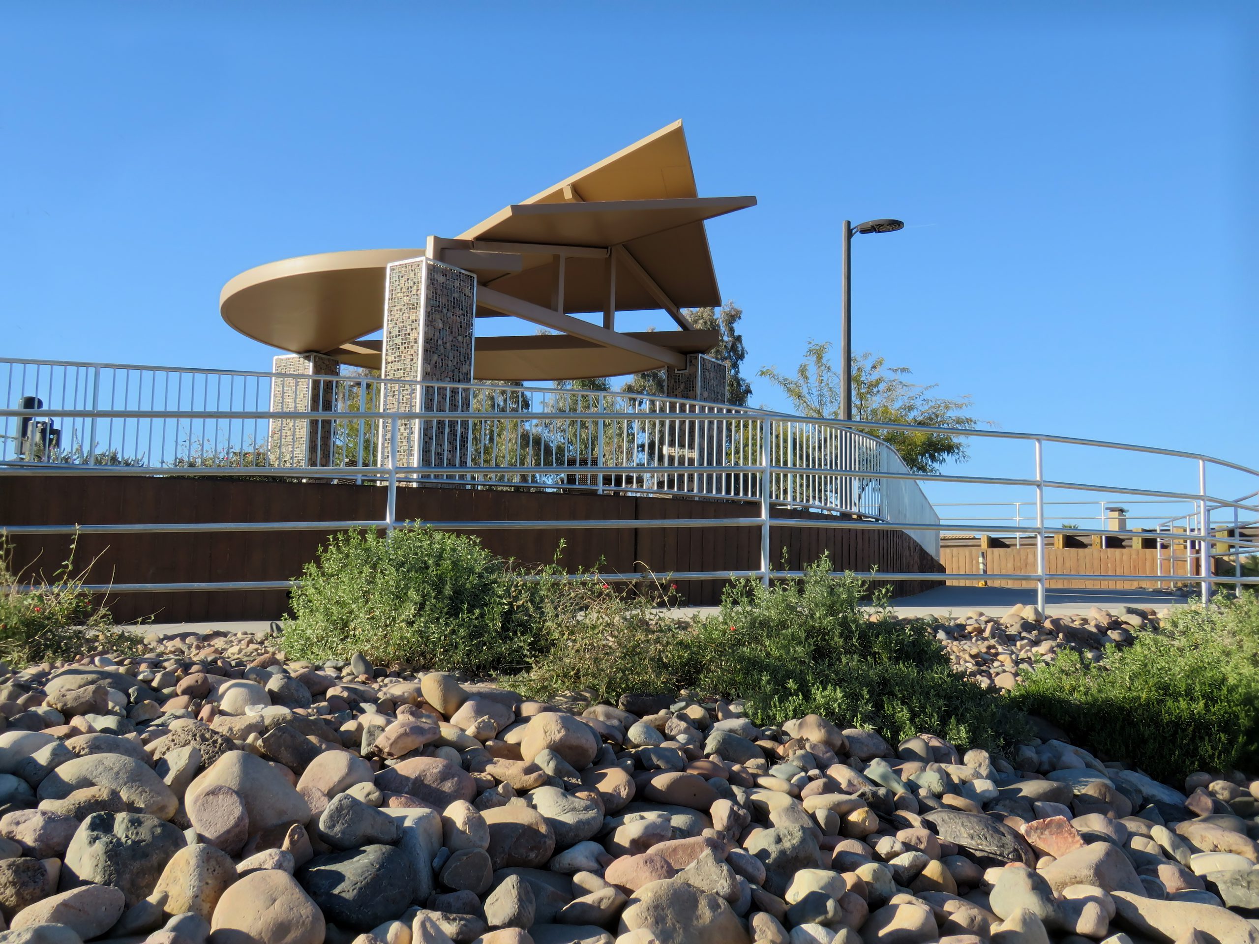 The sculptural shade structure rises up in the background. An accessible ramp in the foreground sweeps through native shrubs and river rock.