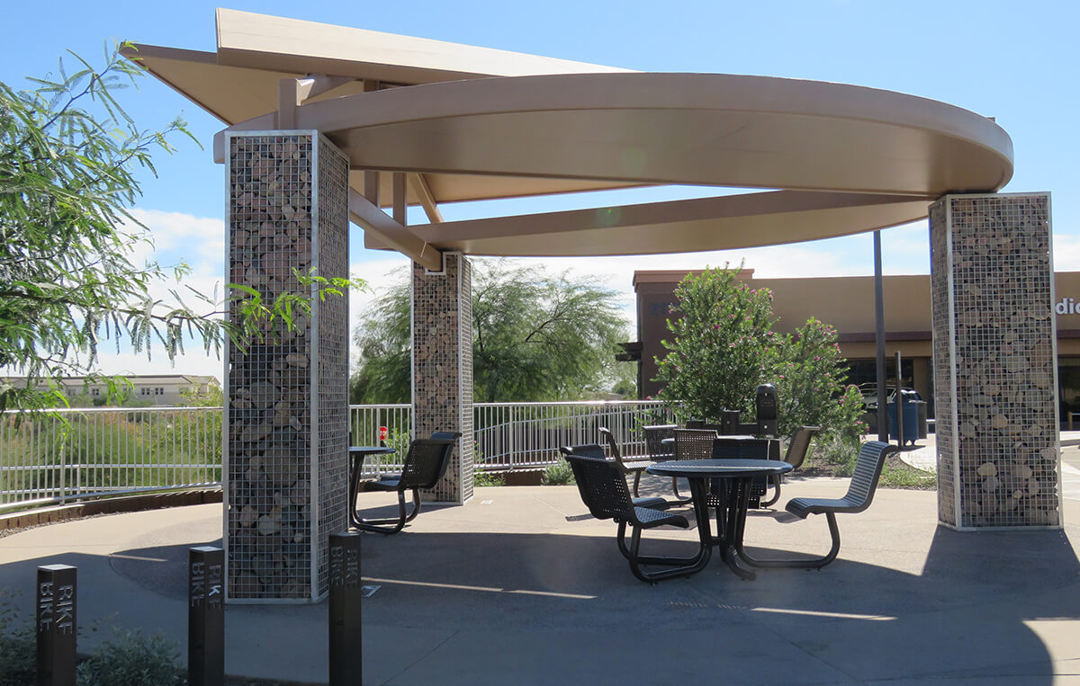 Photo of a shade structure over built-in metal seating on a smooth circular patio. Thick columns made of gabion baskets filled with rock are holding up a circular metal roof.